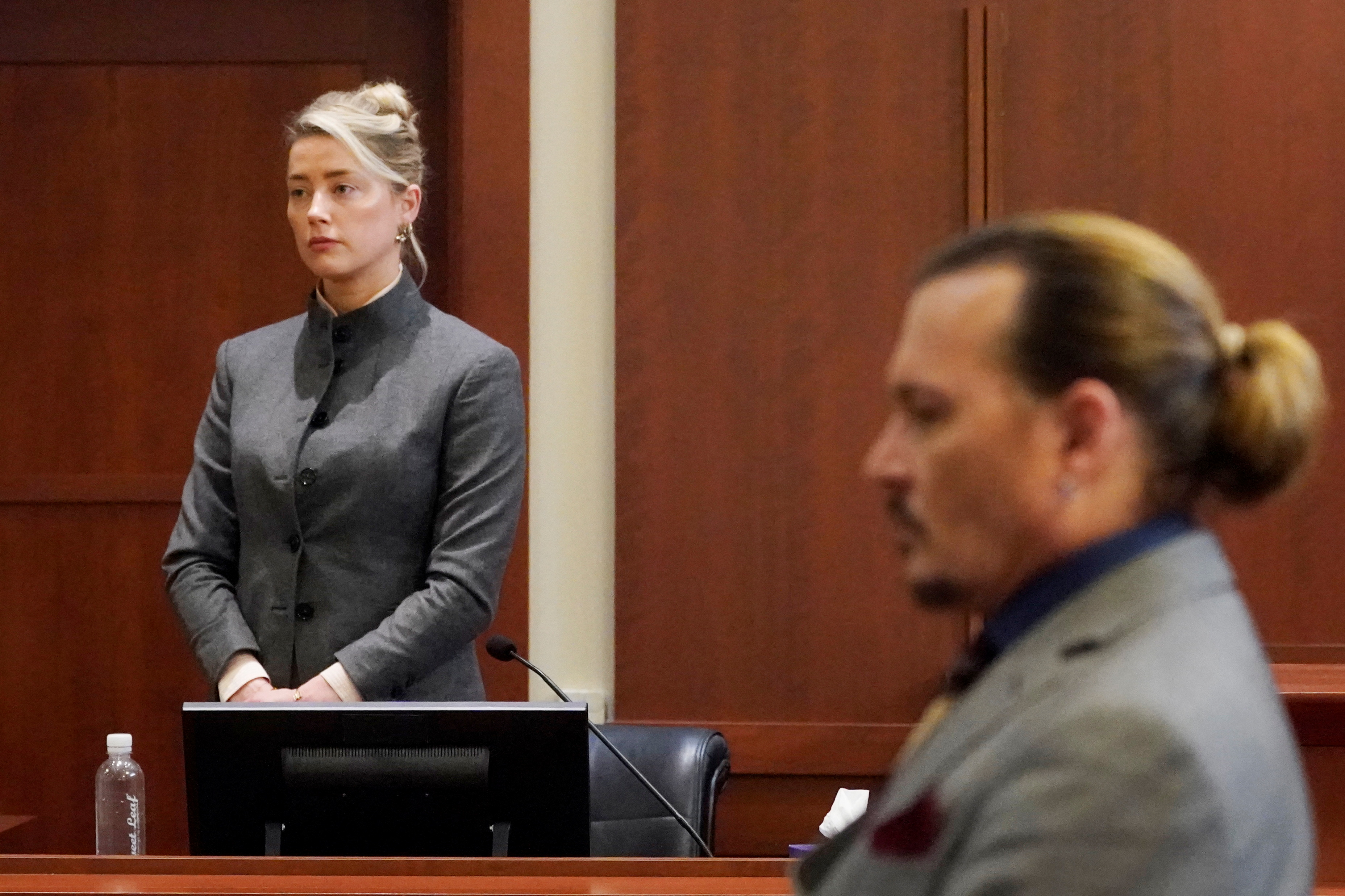 Amber Heard 穿着灰色西装站在证人席上，而 Johnny De pp坐在前台out of foxu